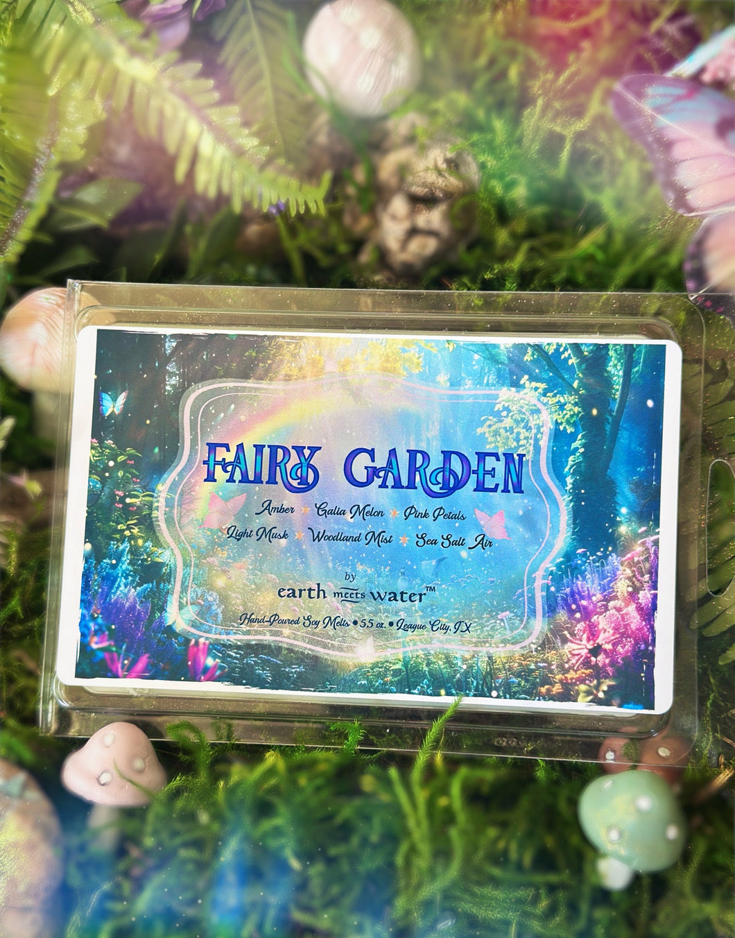 A packaged wax melt or scented melt block labeled 'FAIRY GARDEN' by earth & water, shown in clear plastic packaging with a colorful underwater fairy-garden style label, placed among moss and small decorative stones.