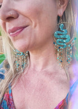 Close-up of a person wearing turquoise snake-shaped earrings with chain fringes.